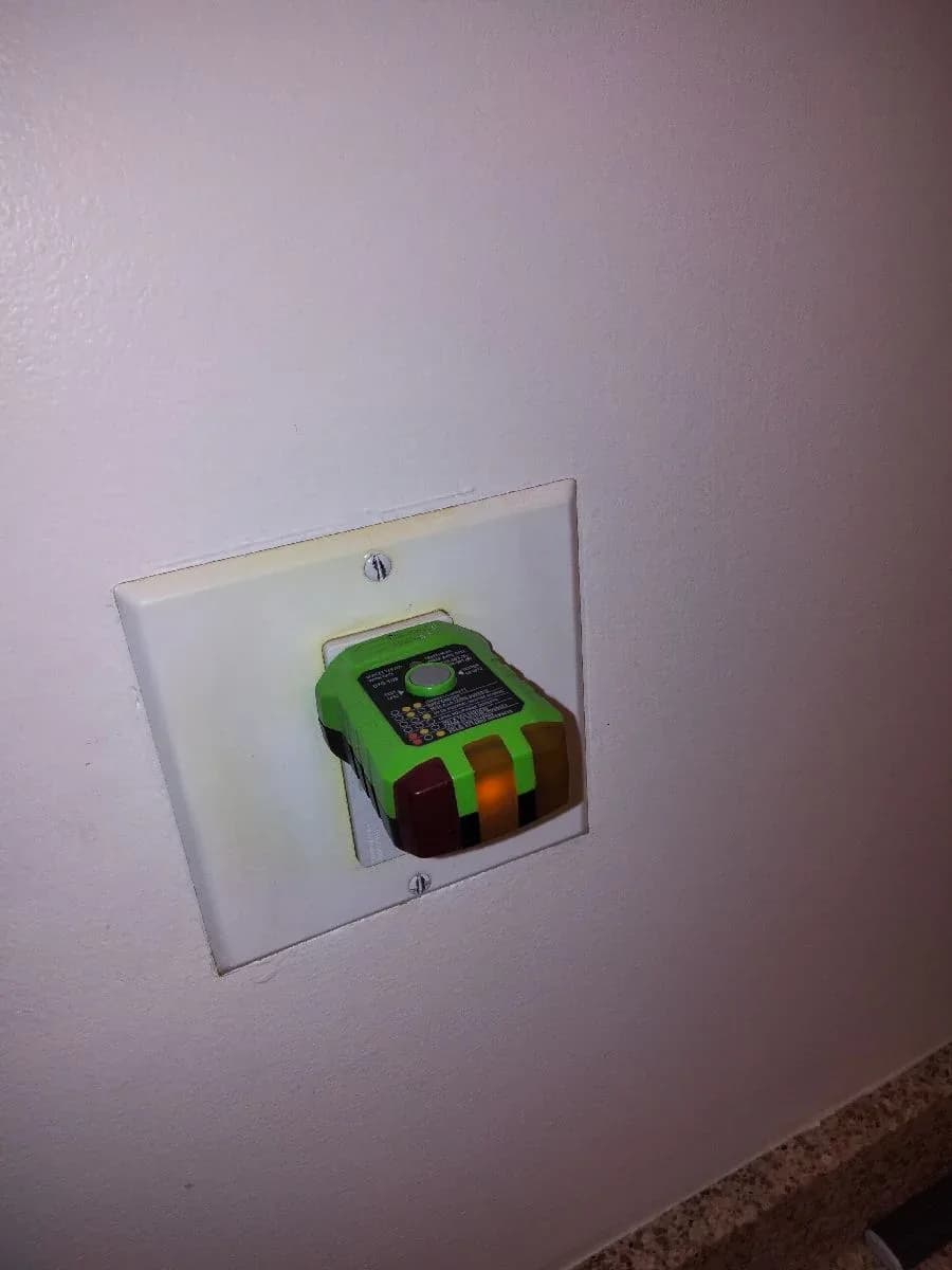 White wall receptacle with a green plug-in electrical tester installed in the lower outlet.
