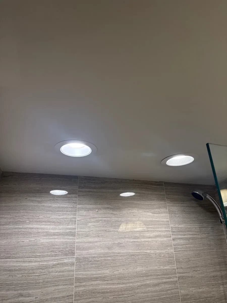 Two illuminated recessed ceiling lights installed above a tiled bathroom wall, with a shower fixture visible at the right.