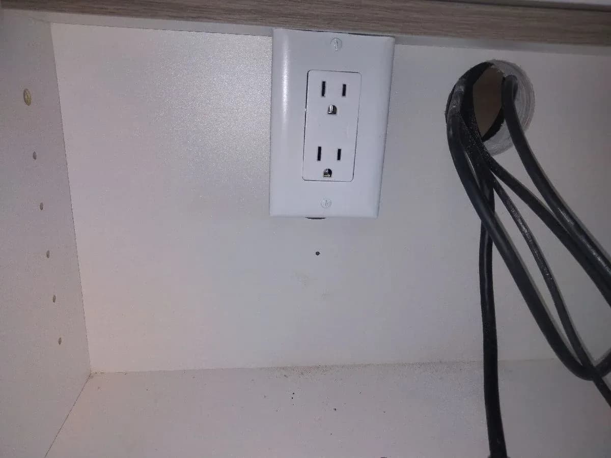 White duplex electrical receptacle installed on the back panel of a cabinet, with black cables routed through an opening to the right.