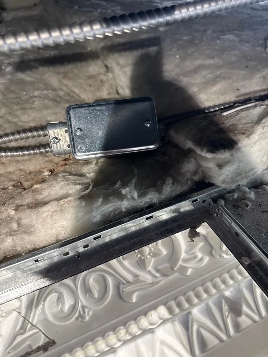 Covered black electrical junction box connected to flexible metal conduit above a suspended ceiling panel.