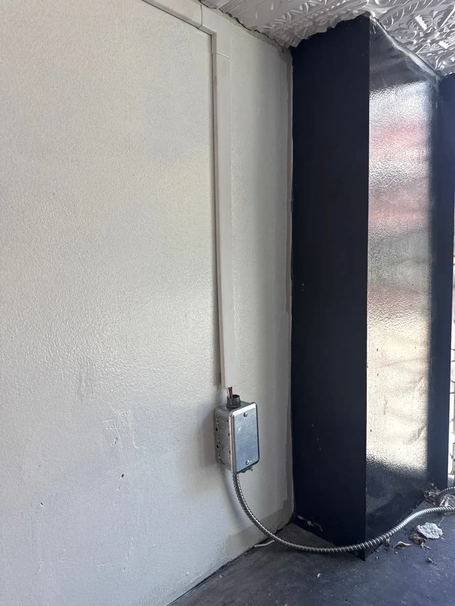 Wall-mounted metal electrical box connected to surface conduit beside a dark vertical panel.