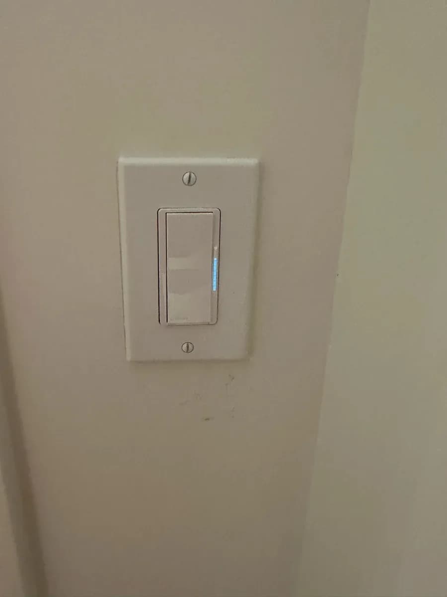 Wall-mounted white electrical dimmer switch with a small blue indicator light on a beige wall.