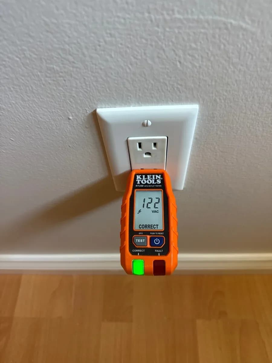 White wall outlet with an orange plug-in electrical tester displaying 122 VAC and a correct wiring indication.
