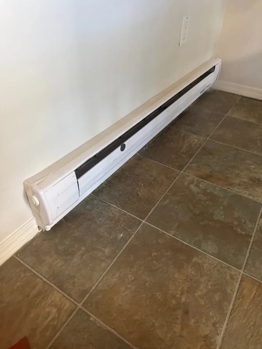 White electric baseboard heater installed along a light-colored wall above brown tiled flooring.