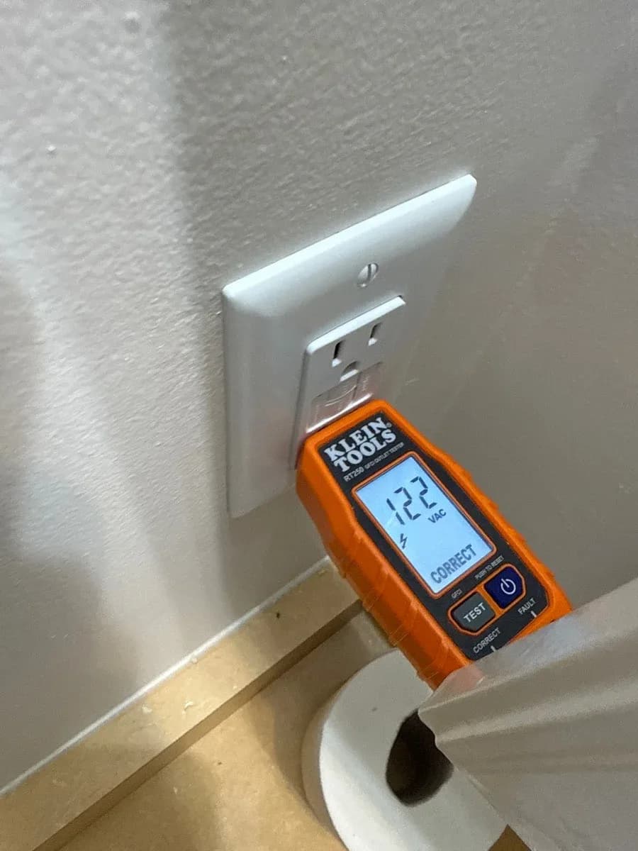 White wall receptacle being tested with an orange plug-in electrical tester displaying 122 VAC and a correct indication.