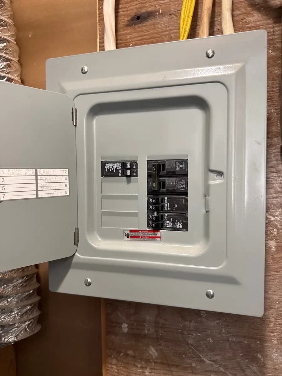 Open gray electrical breaker panel with several installed circuit breakers, handwritten circuit labels, and insulated cables visible around the enclosure.