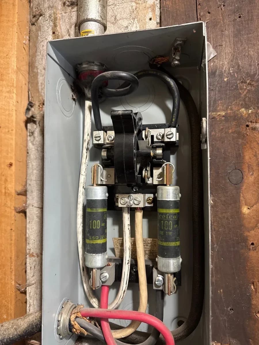 Open gray electrical service enclosure showing two cylindrical 100-amp fuses, wiring, terminals, and conduit.