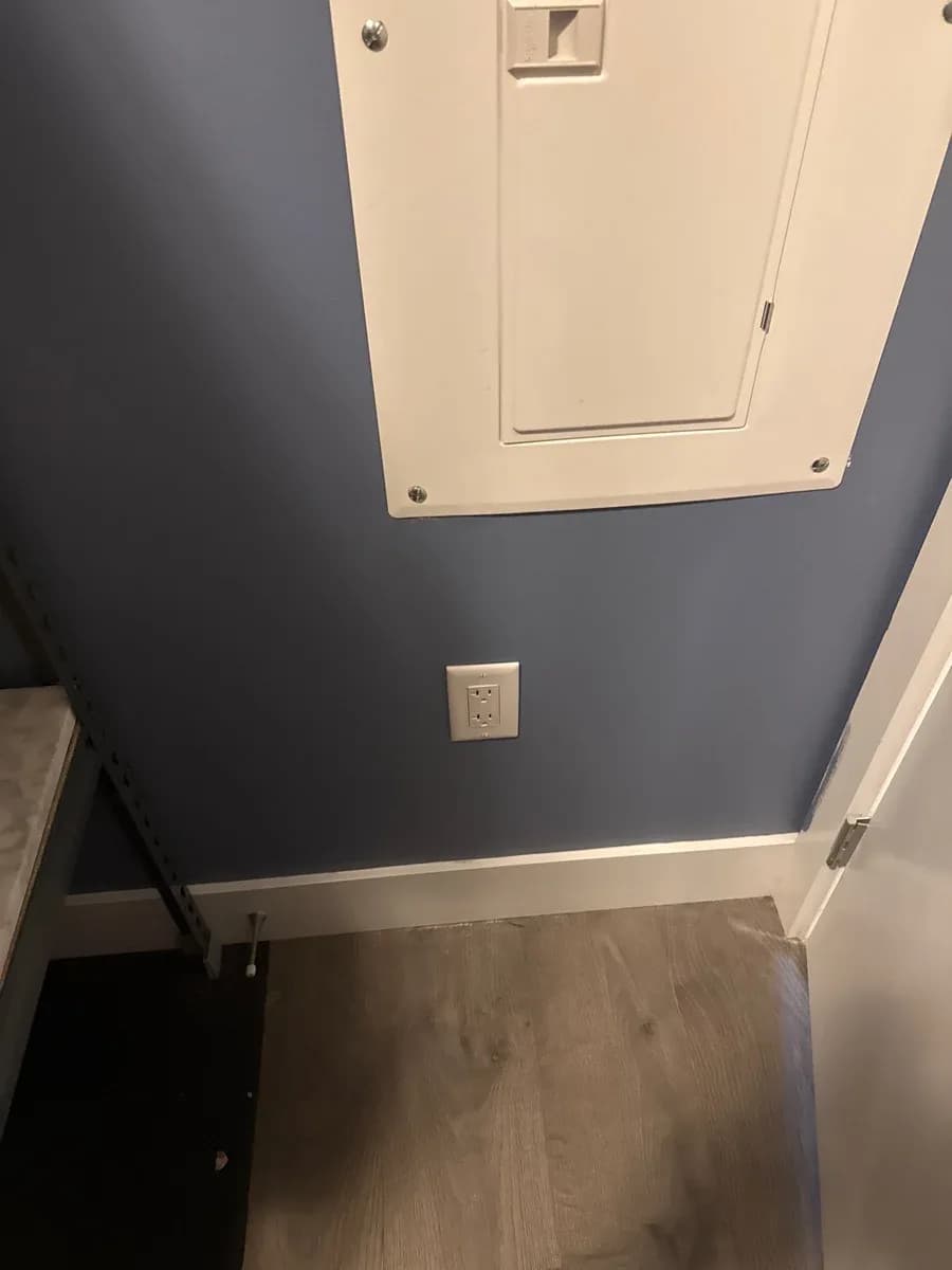 Closed white electrical panel and duplex receptacle installed on a dark blue wall above a wood-look floor.