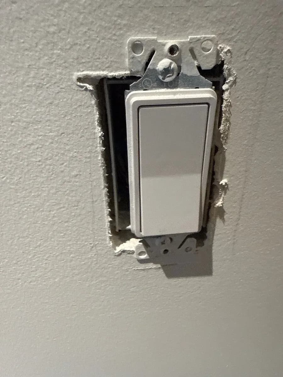 White electrical wall switch installed in a gray textured wall, with the wall plate and surrounding drywall unfinished.