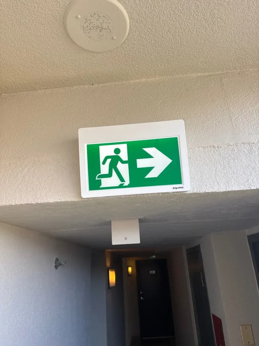 Building corridor with a green illuminated exit sign pointing right, a ceiling-mounted detector, and a wall-mounted emergency light.