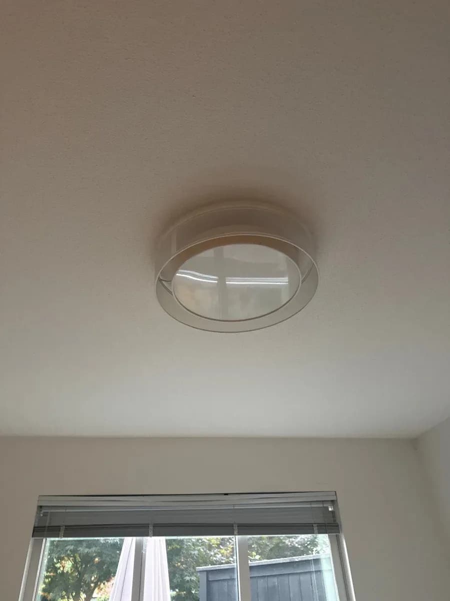 Flush-mount round ceiling light fixture installed on a white ceiling above a window with horizontal blinds.
