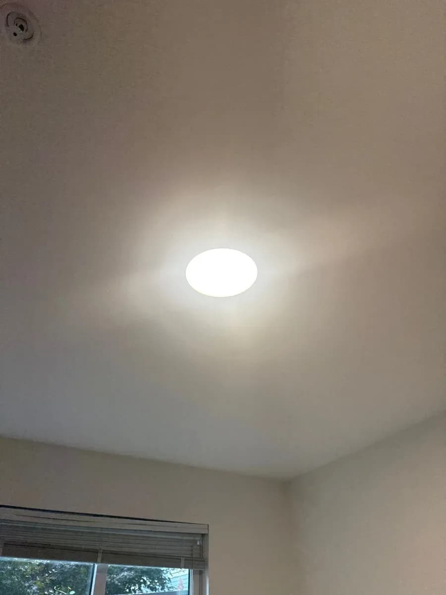 White circular recessed ceiling light illuminated in a finished room, with a window and blinds visible below.
