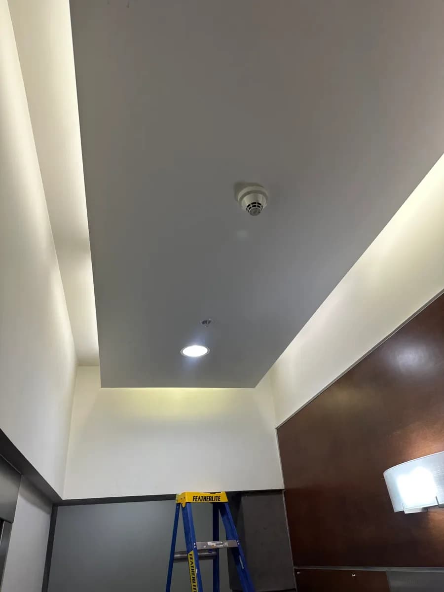 Interior ceiling and wall showing a lit recessed light, ceiling-mounted detector, wall light, and a ladder below.