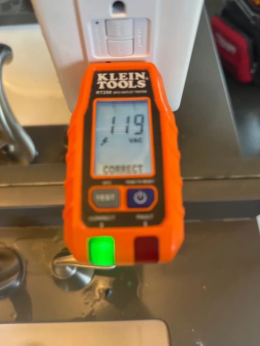 Klein Tools voltage tester displaying 119 VAC at a white electrical receptacle above a countertop.