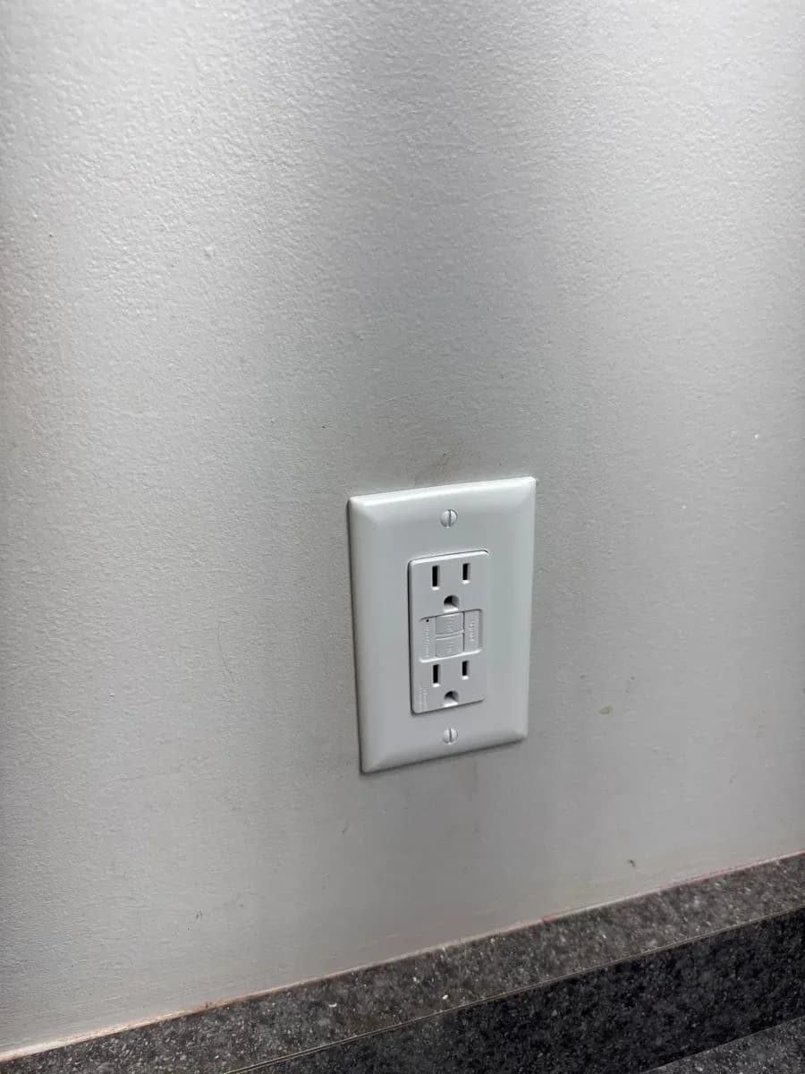 White GFCI electrical receptacle with a matching wall plate installed on a light-colored wall above a dark baseboard.