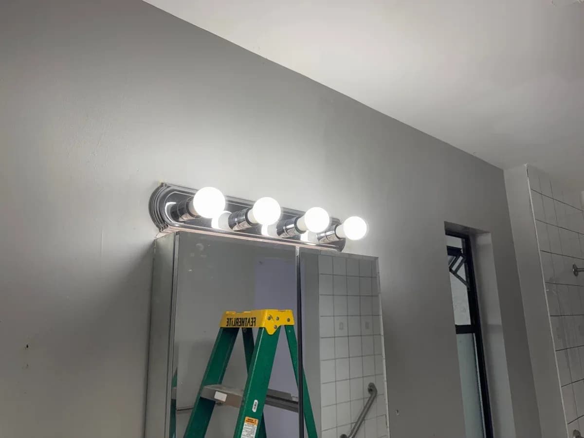 Illuminated four-bulb vanity light installed above a bathroom mirror, with tiled walls and a green step ladder reflected in the mirror.