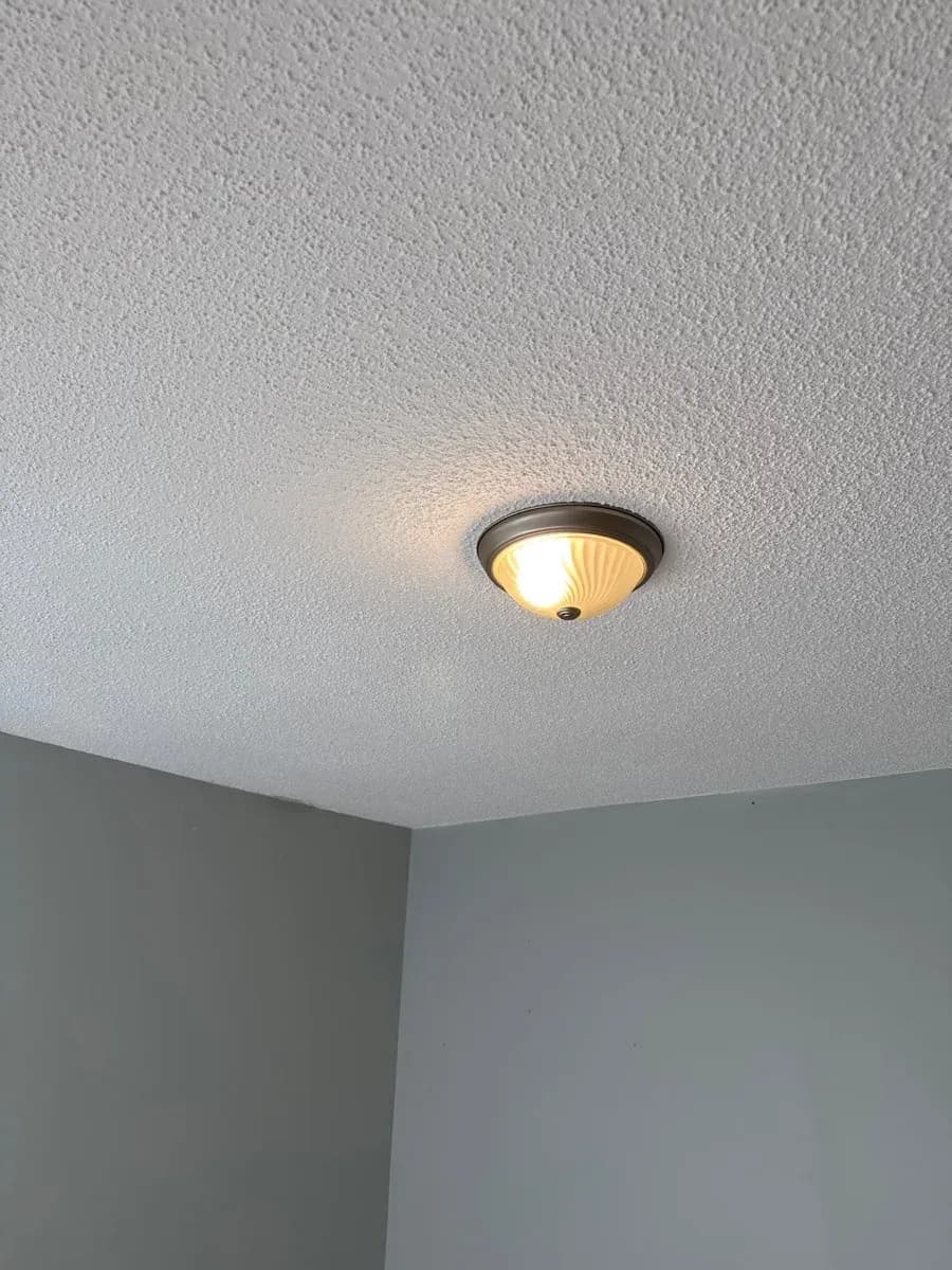 Illuminated flush-mount ceiling light fixture installed on a textured ceiling above a gray-walled room corner.