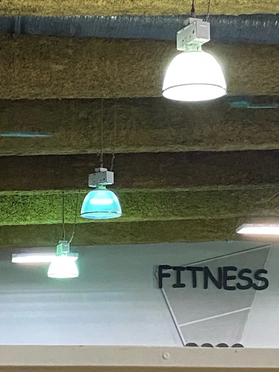 Several illuminated pendant and linear light fixtures installed beneath exposed ceiling beams in an indoor fitness area.